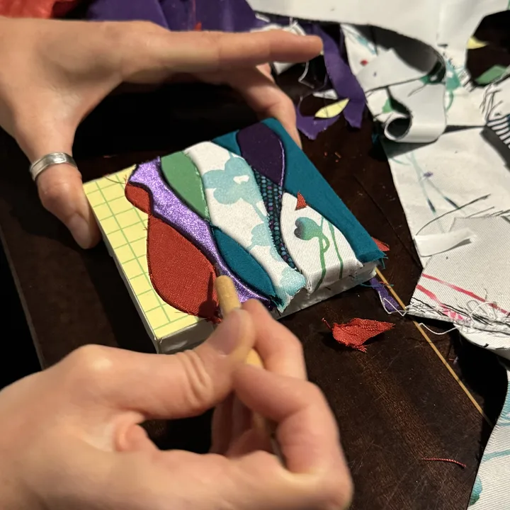 Mains d'une participante incrustant des tissus colorés dans son tableau, pendant un atelier