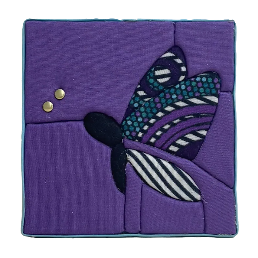 Tableau textile Kinusaiga de 10 × 10 cm représentant un papillon : corps noir, ailes en wax à motifs violets, turquoise, noirs et blancs, sur fond violet, avec deux petits rivets dorés au bout des antennes, bordé d'un biais bleu ciel.