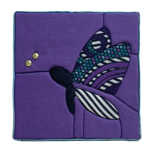 Tableau textile Kinusaiga de 10 × 10 cm représentant un papillon : corps noir, ailes en wax à motifs violets, turquoise, noirs et blancs, sur fond violet, avec deux petits rivets dorés au bout des antennes, bordé d'un biais bleu ciel.